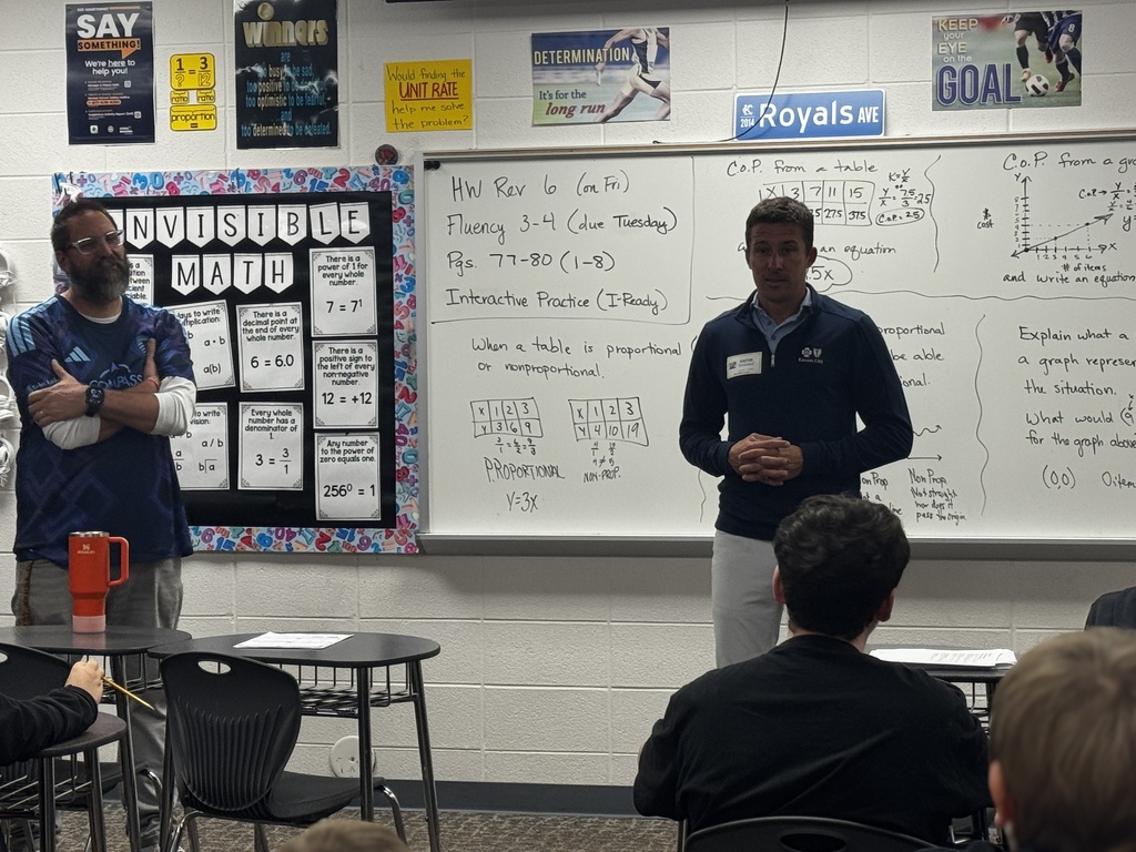 Two men in blue shirts stand in front of a classroom. A whiteboard with writing and a bulletin board are in the background.