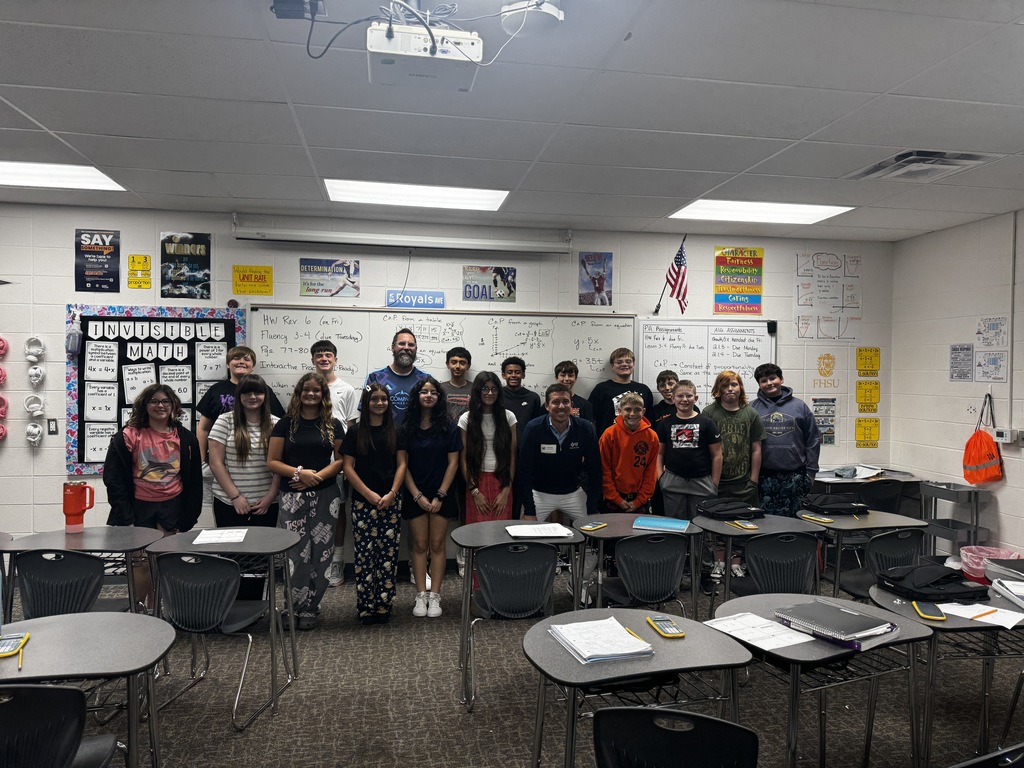 A group of students and teachers are in a classroom with desks and tables. There are posters on the walls.