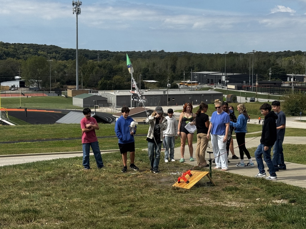 People gather in a park, with some observing a rocket launch in the distance.