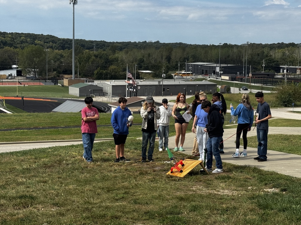 Group of people standing on a grassy field, engaging in a game of cornhole.
