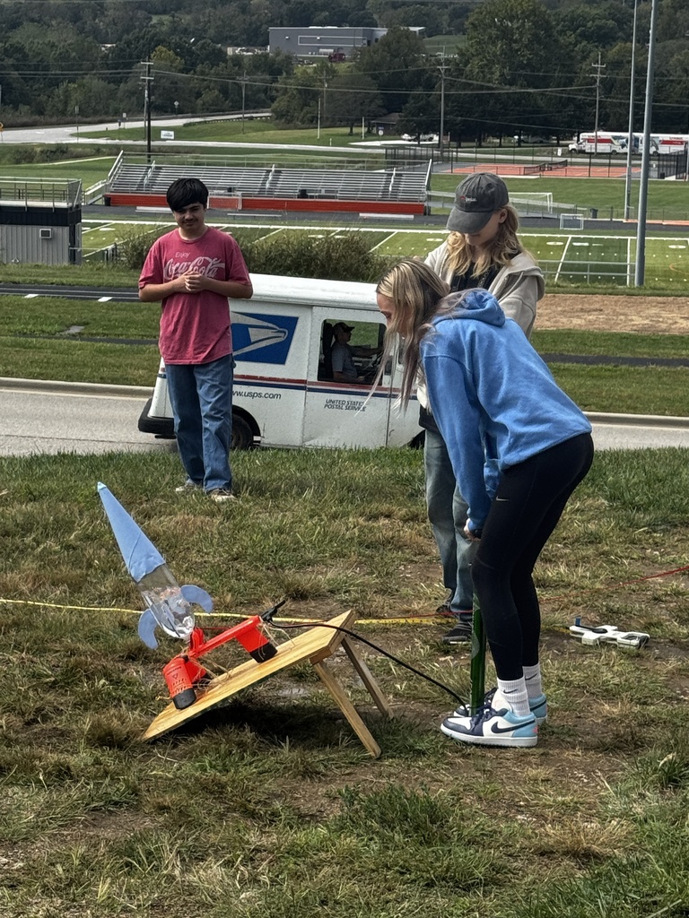 Three people on a grassy field with a rocket launcher. The rocket is on a stand.