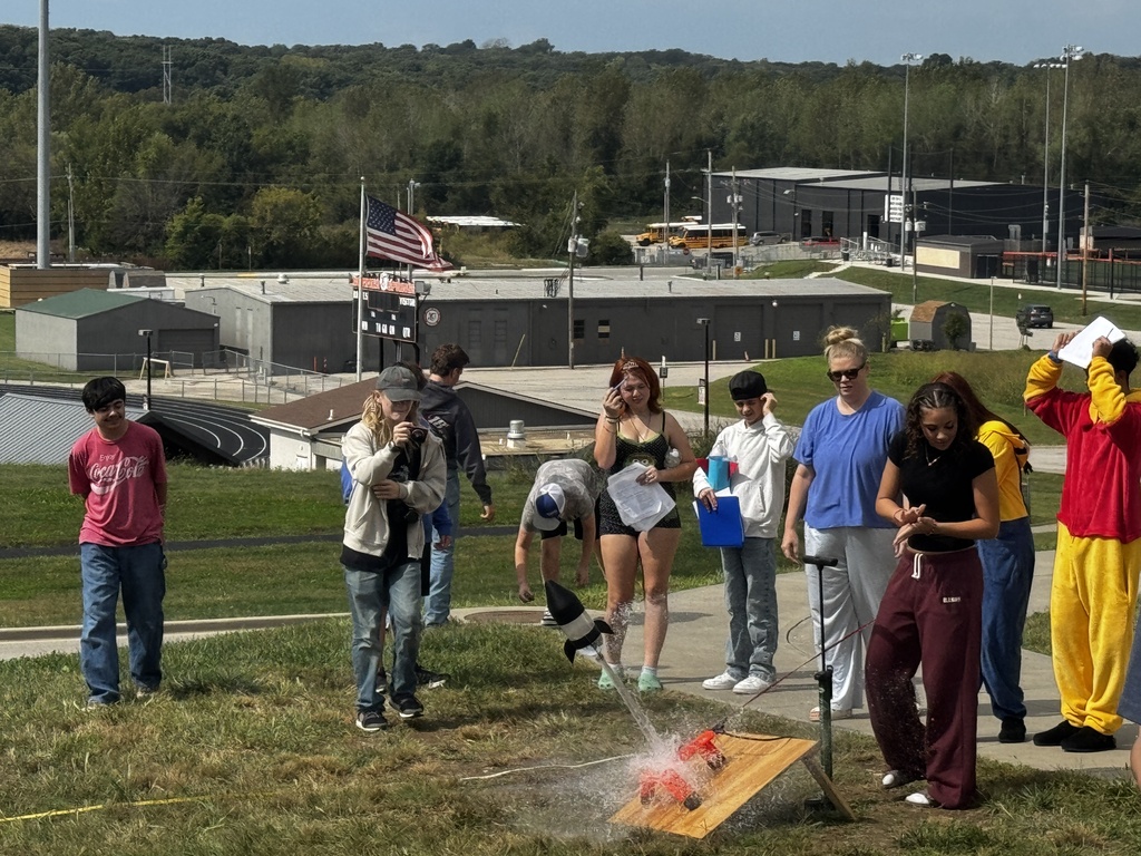 Group of people gather around a science experiment on grass, with a flag and buildings in the background.