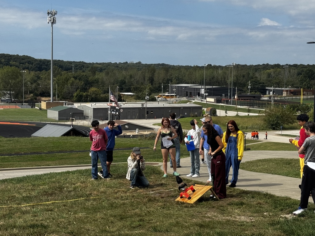 Group of people outside on grass near a cornhole board. Background features trees and buildings.