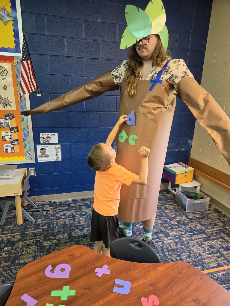 A child in a yellow shirt stands with arms up near a teacher in a brown costume, indoors.