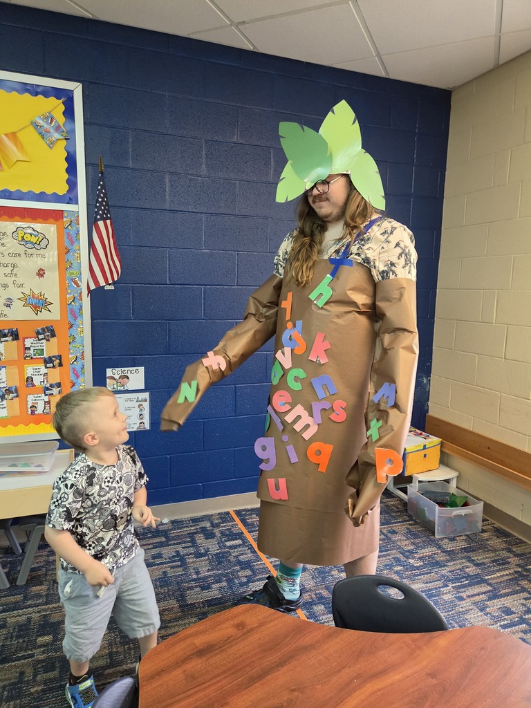 A person dressed as a giant tree interacts with a child in a classroom with blue walls and a flag.