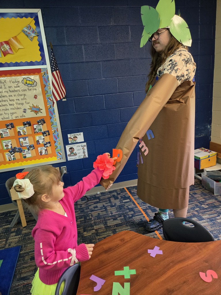 Girl in a pink shirt and green skirt holding flowers in a classroom. A woman in a brown dress and green hat stands nearby.