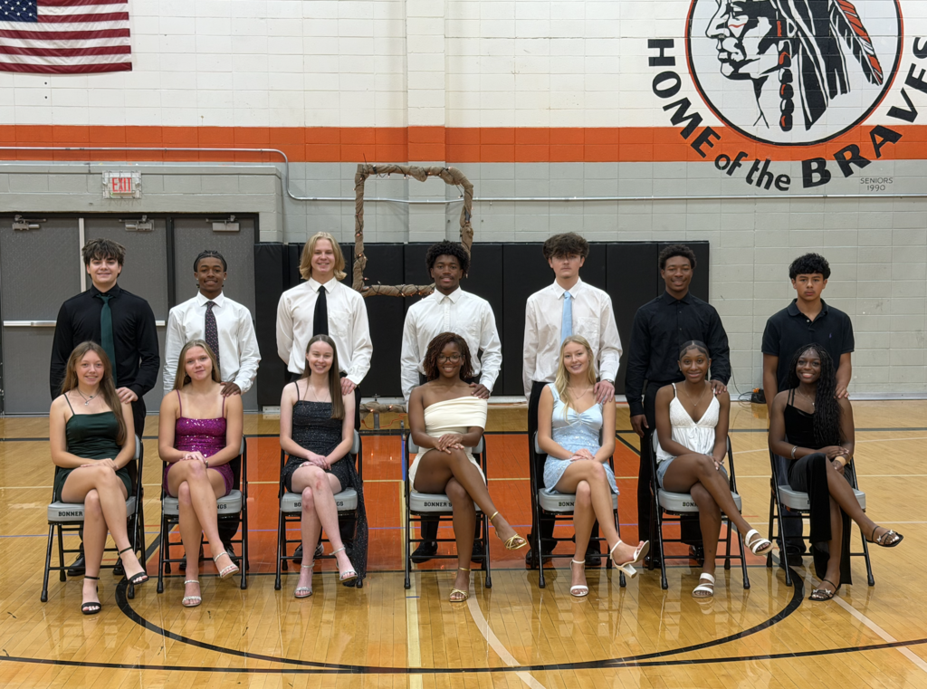 A group of students in formal wear seated in chairs on a basketball court. The background includes a school logo.