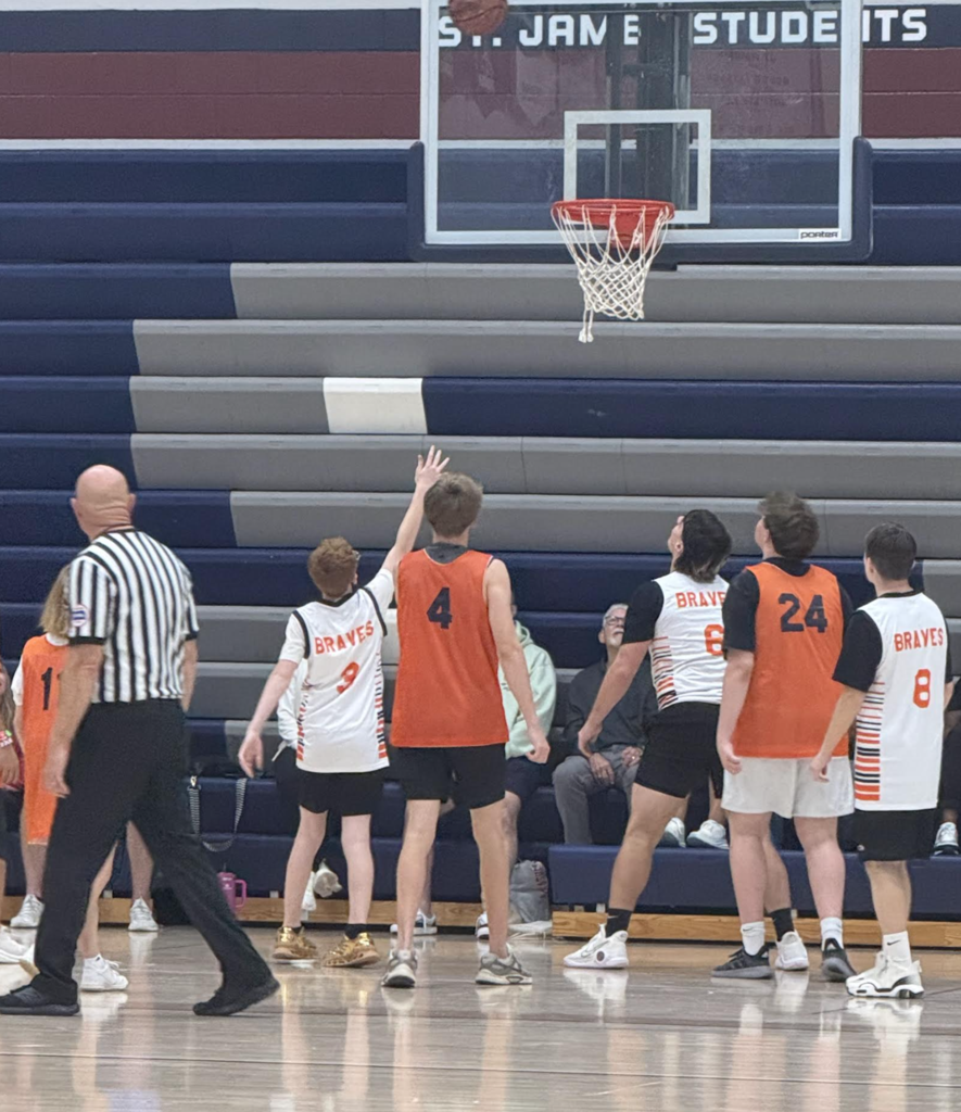 Indoor court with players in numbered jerseys, a referee walking, and a basketball hoop. Audience seated in stands.
