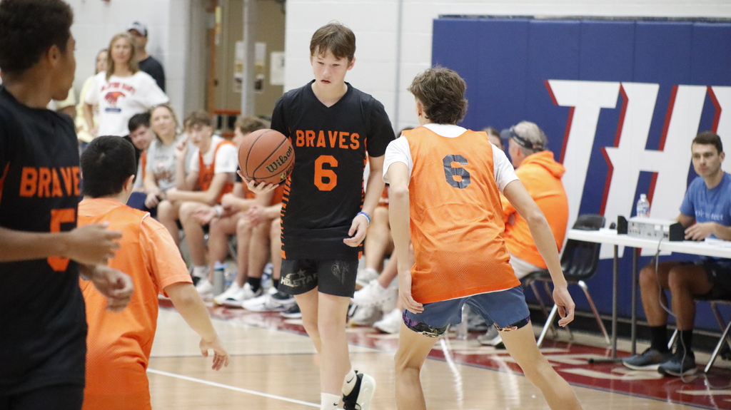 Players in jerseys are on a basketball court. One boy holds a ball. Spectators are seated in the background.