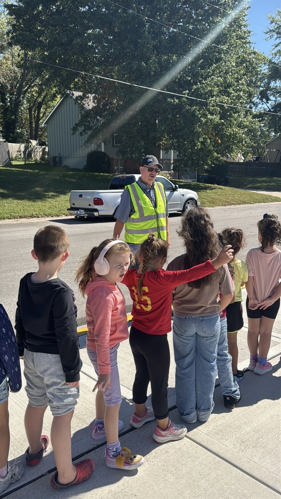 Adults and children stand in a line on the sidewalk. One adult wears a high-visibility vest. A pickup truck drives past them.