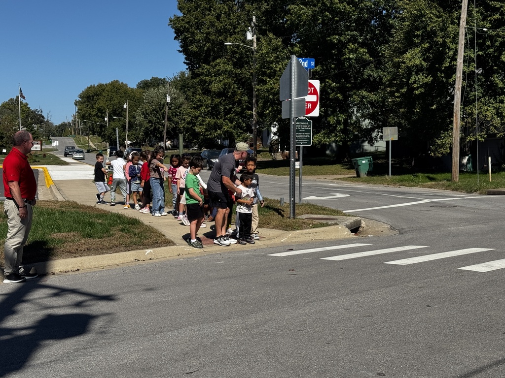 People stand at a crosswalk. A man in a red shirt stands on the sidewalk. A traffic sign is nearby.
