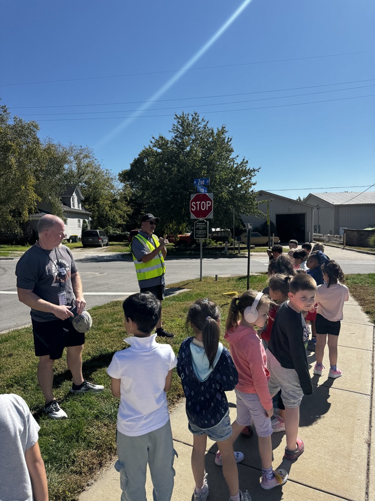 A group of children waits at a stop sign, accompanied by a man, with a stop sign and buildings in the background.
