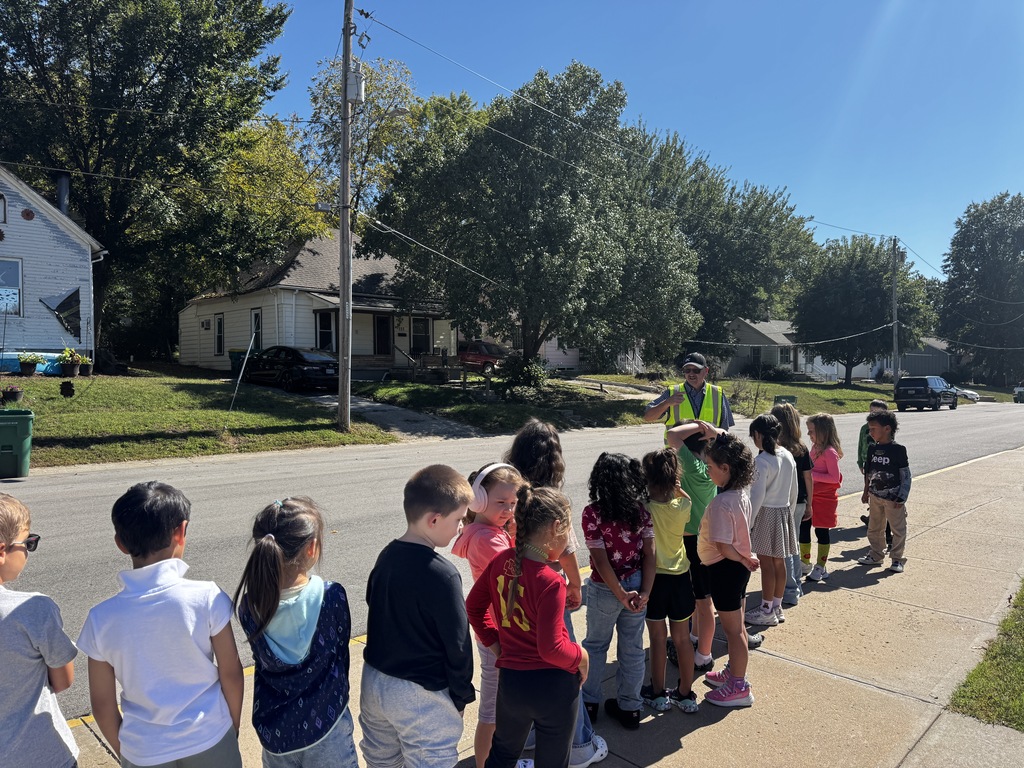 A group of children listens to a person in a safety vest on a street with houses and trees.
