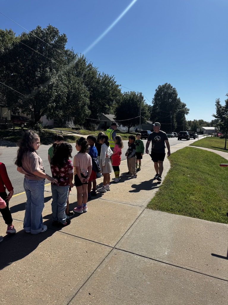 Children and an adult standing on a sidewalk under a blue sky, with trees and grass.