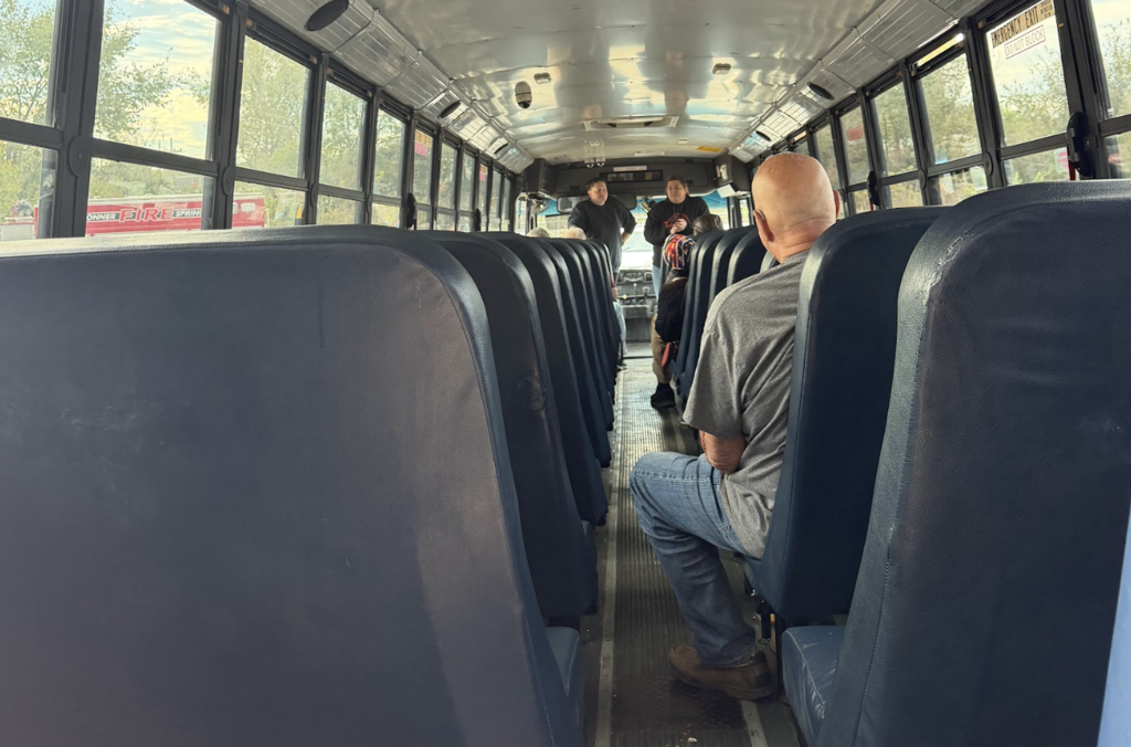 Interior of school bus with rows of blue seats; man sitting and another standing near the back.