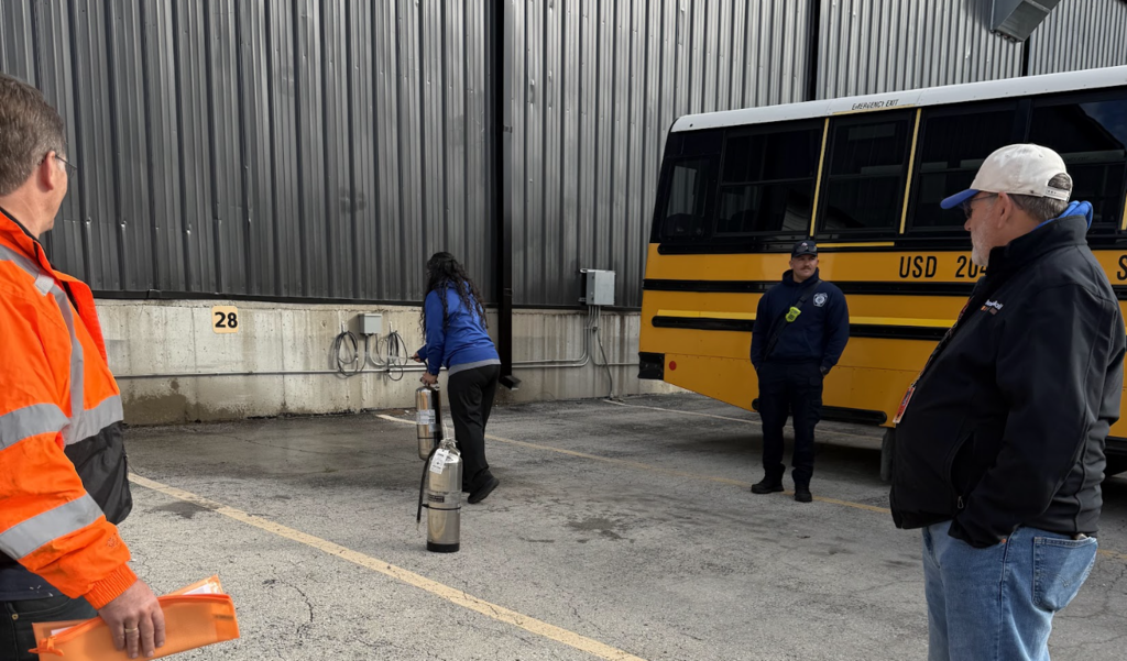 Four adults, one holding a folder, stand by a yellow bus with a large silver tank on the pavement.