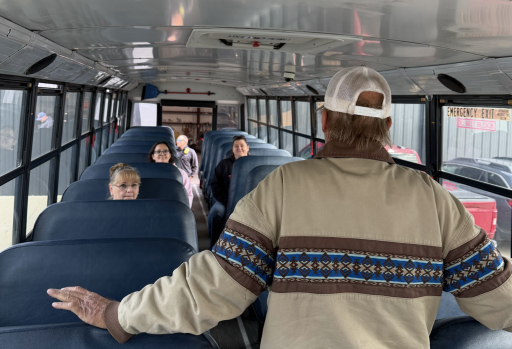 A man faces away from seated children on a school bus, hand extended. He wears a tan jacket.