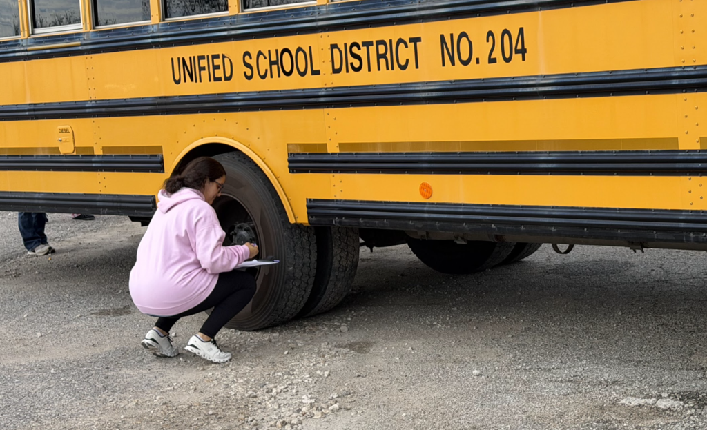 A student crouches next to a school bus tire, holding a pen and paper.