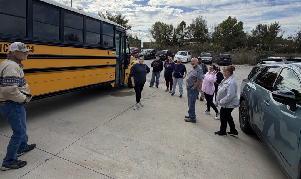 People stand near a school bus. Some people walk between vehicles. A parking lot with cars is in the background.