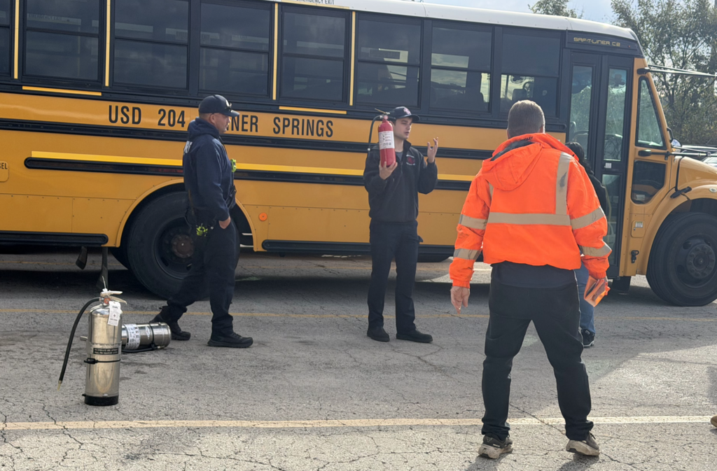 Three people in jackets and hats stand in front of a school bus. One holds a fire extinguisher.