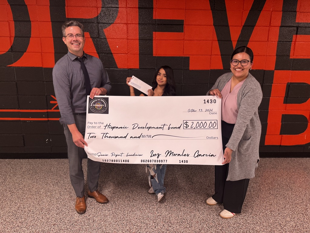 Three people hold a large check over their heads in front of a red brick wall.