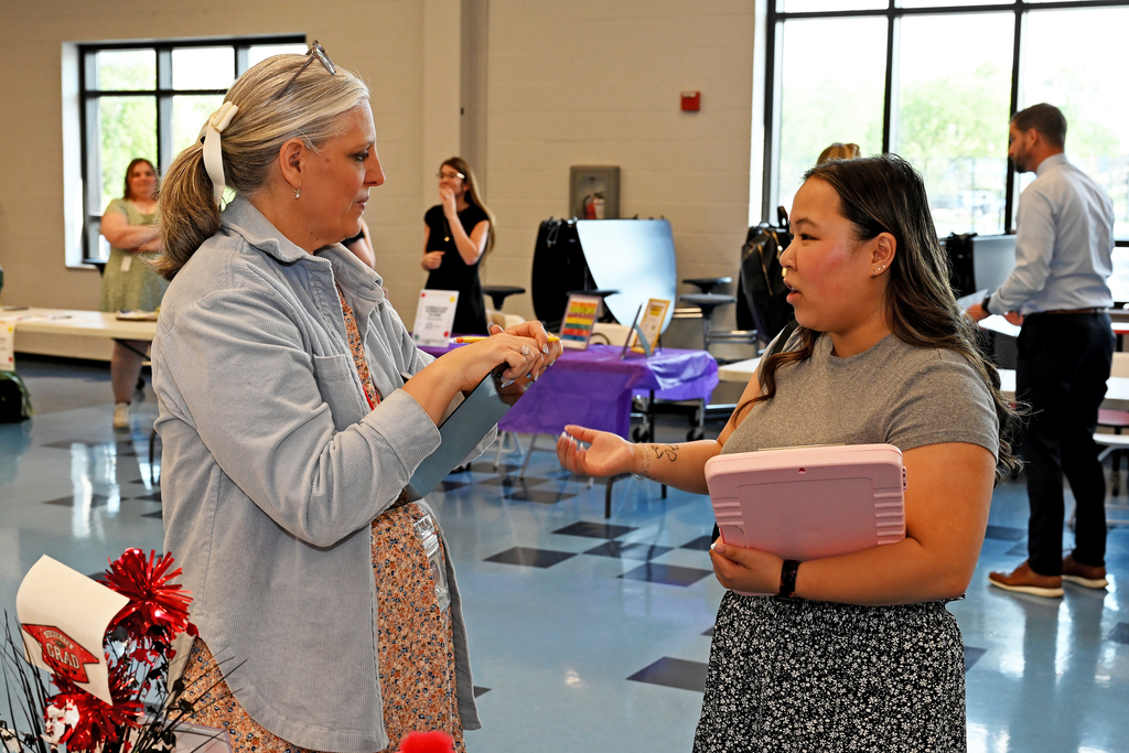 Two women face each other in a room, one holding a pink folder, the other gesturing. Behind them, people stand at tables with various items.