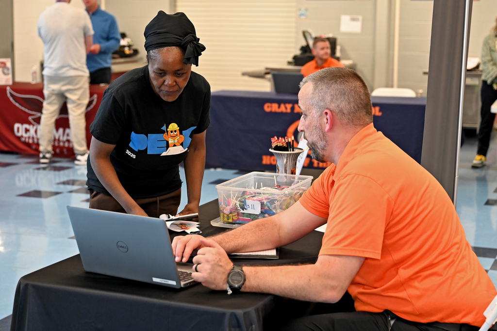 A man in an orange shirt at a table uses a laptop while a woman in a black shirt looks on.