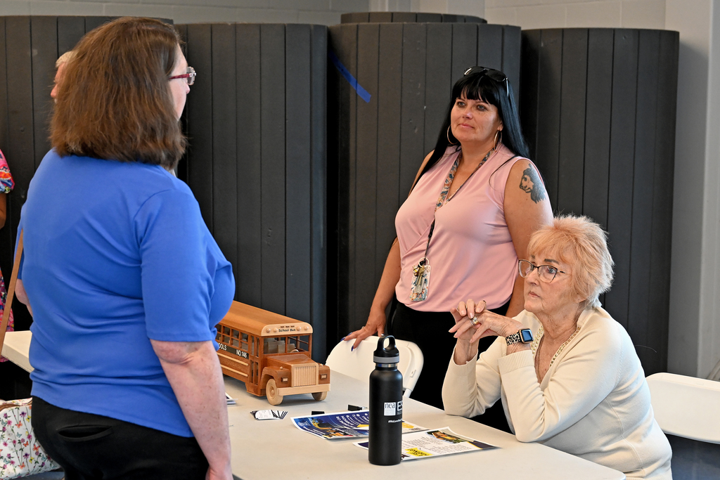 Three women are gathered around a table, with one woman in blue standing and speaking to another woman in white.