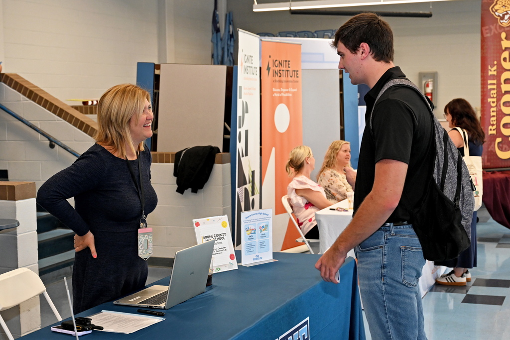 Two people in a room with a table, one holding a laptop and papers, talking to a man with a backpack.