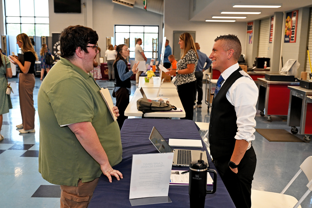Two people stand at a table in a room, conversing. One man wears glasses and a green shirt. A woman wears a vest and white shirt.