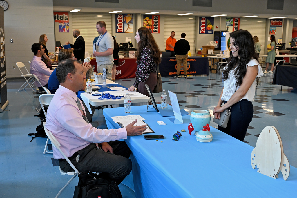 People in a room with blue floors and white walls. A man sits at a table with another person standing.