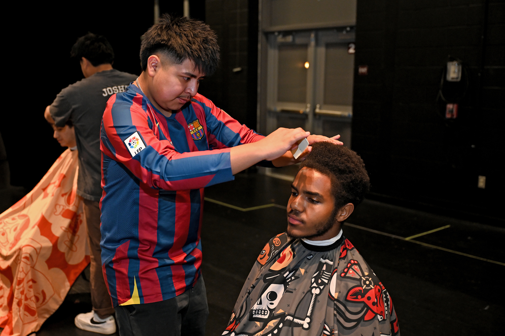 A man in a striped shirt cuts another man's hair in a room with a black wall.