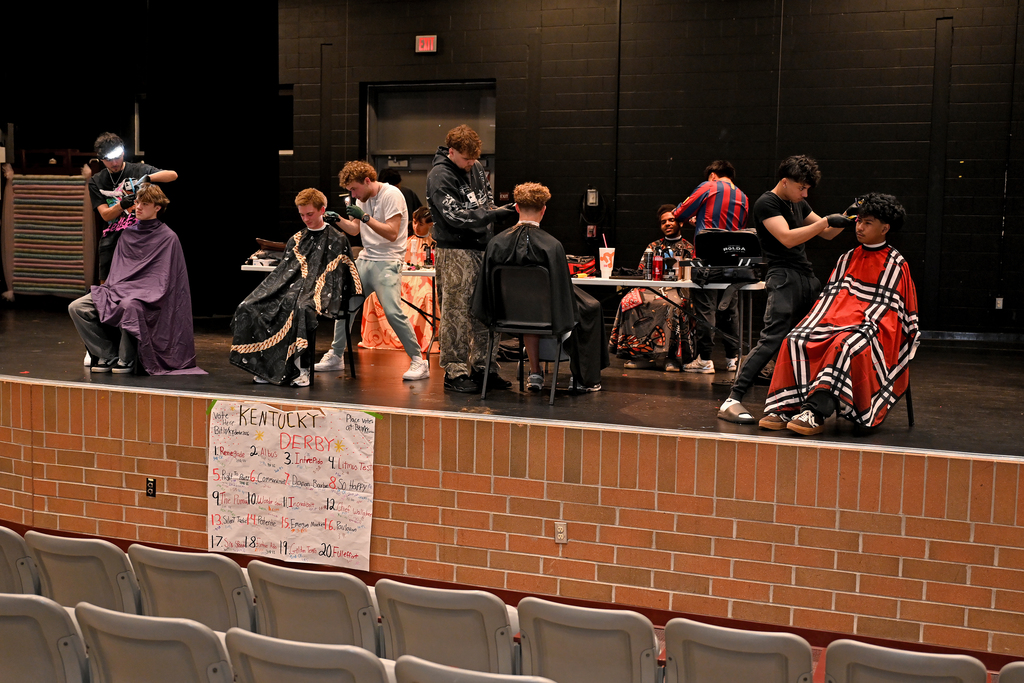 People on stage, some sitting, some standing, with chairs and tables; several getting haircuts.