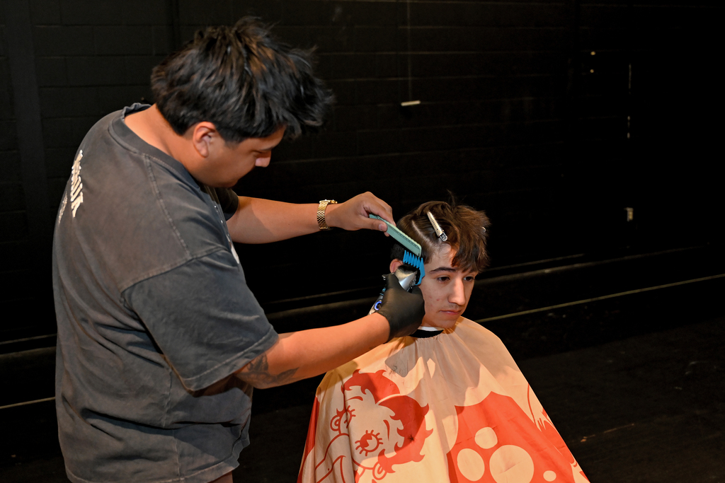 A person in a gray t-shirt cuts another person's hair using clippers in a dimly lit room.