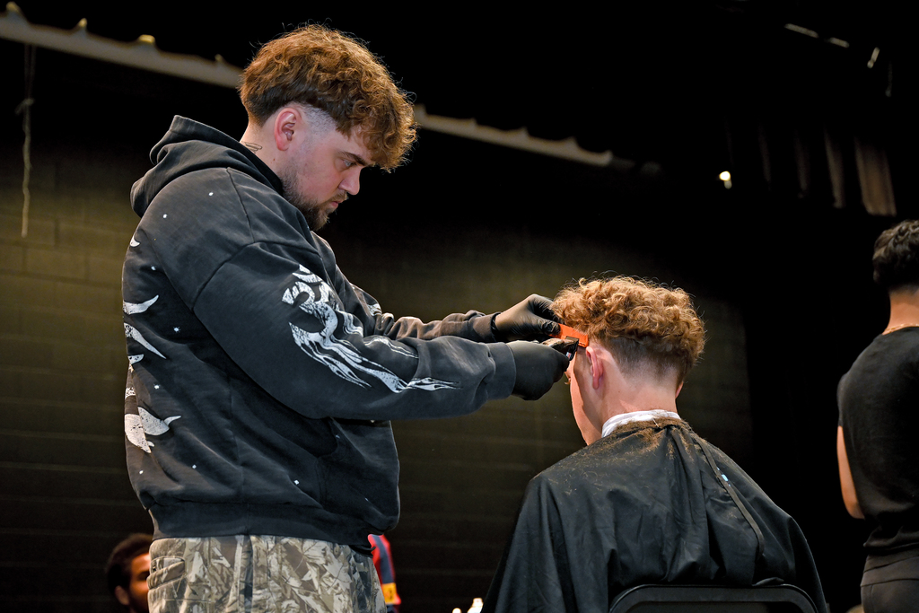 A man in a hoodie, standing, cuts hair of a seated person. Another person stands nearby.