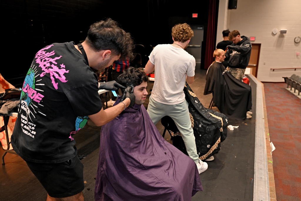 A person cuts another's hair in a theater, with two others getting their hair done in the background.
