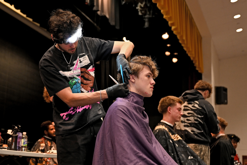 A man cuts another man's hair with a scissor in a room with stage backdrops and a table.