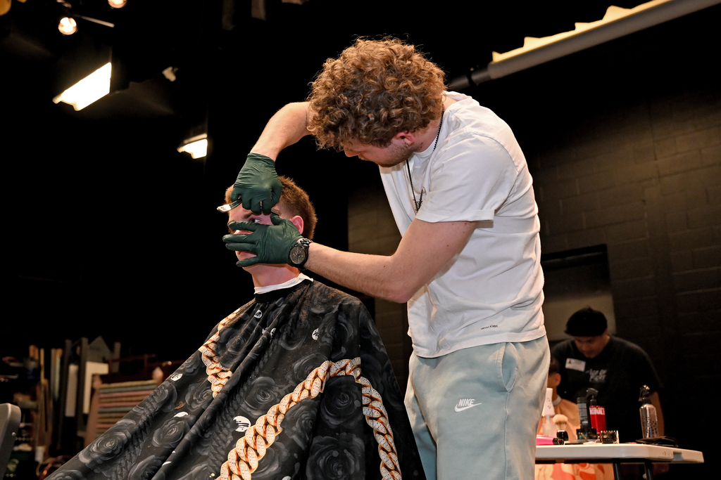 Man with curly hair standing cuts hair of a seated person wearing a black cape.