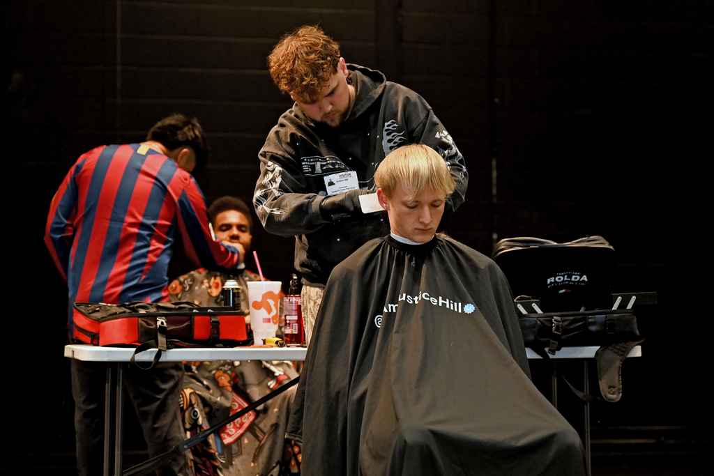 Three people are on a stage. One sits in a chair, getting a haircut. Another stands behind them. 