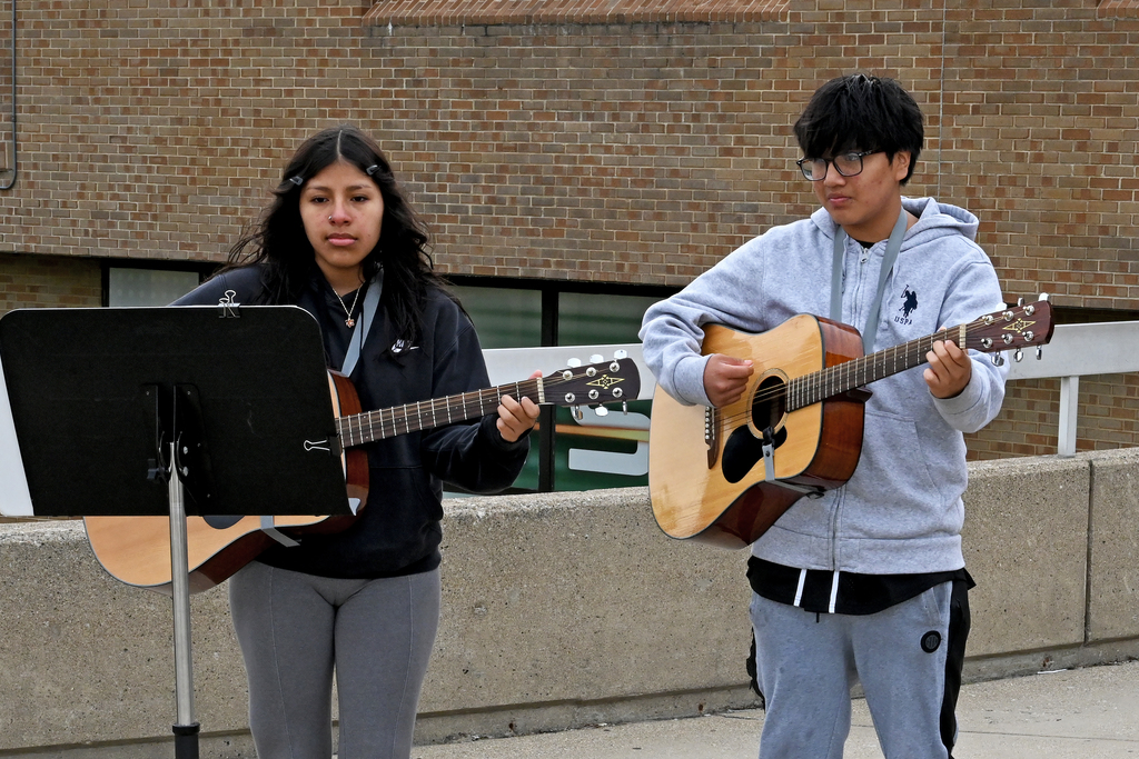 Two individuals stand on a concrete platform, each playing a guitar, with a music stand in front of one.