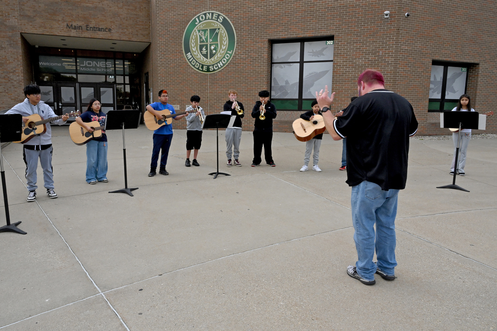 Several individuals play guitars and wind instruments outdoors in a circle, led by a conductor 