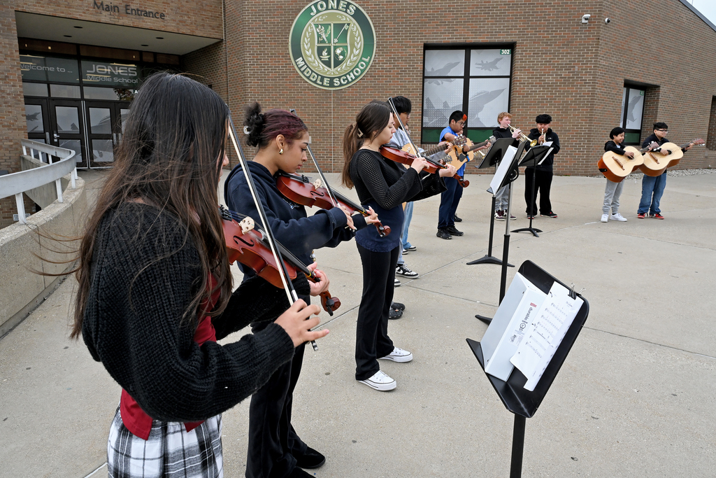 A group of students plays stringed instruments on a concrete platform in front of a school building.