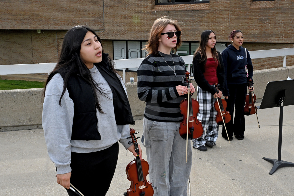 Four women standing in front of a building, one holding a violin and bow, two with violins, one with a music stand.