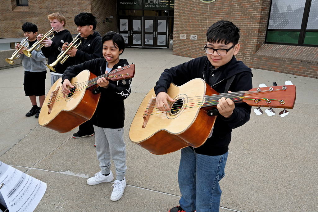 Five children, two playing guitars and three playing trumpets, stand in front of a building. They are smiling and appear to be performing.
