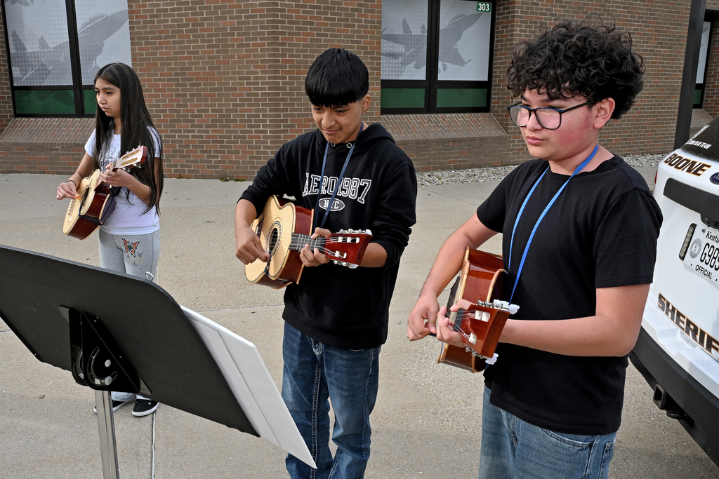Three students in school uniforms play guitars outside a brick building, with a music stand and a police vehicle.