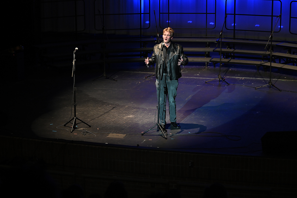 A man stands on stage under a spotlight, holding a microphone, with an empty microphone stand behind him and blue lighting.