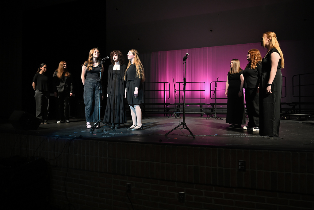 Seven students in black attire on a stage, with one singing into a microphone and pink lighting behind them.