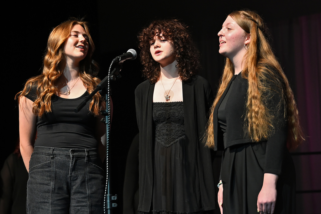 Three students in black clothing sing on stage in front of a microphone. One wears a necklace.