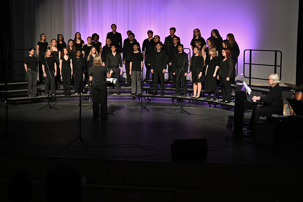 Group of students on a stage, wearing black, with a pianist, a conductor, and purple lighting.