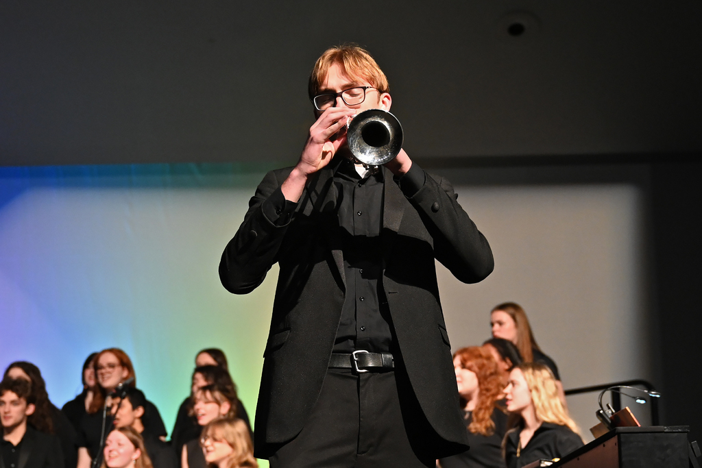A man in a black suit and glasses stands in front of a choir, holding a trumpet to his mouth.
